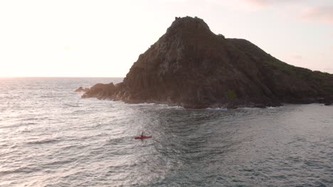 Drone-Aéreo-Dando-Vueltas-Sobre-El-Hombre-Remando-En-Kayak-Rojo-Frente-A-La-Costa-De-Las-Islas-Hawaianas-De-Na-O-Na-Mokulua-Al-Atardecer,-Hawai