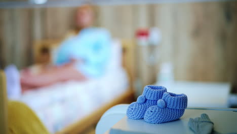 tiny knitted boots for a child expected soon. pregnant woman in maternity hospital ward before labour at backdrop in blur.