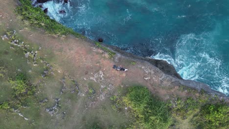 Blue-ocean-waves-break-onto-tall-ocean-cliff,-man-on-moto-sits-on-top