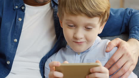 Close-Up-Of-The-Cute-Small-Boy-Sitting-Next-To-His-Good-Looking-Father,-Playing-A-Game-On-The-Smartphone-And-Smiling