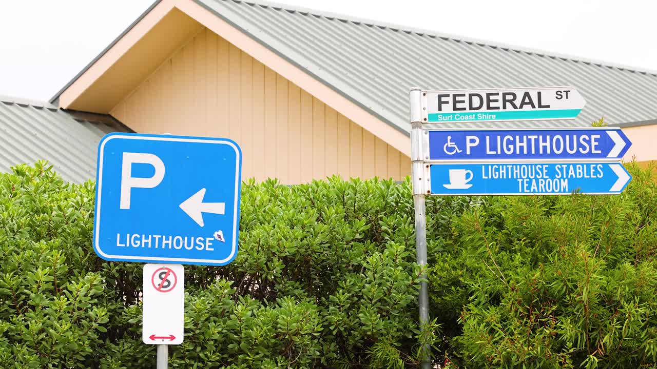 Blue and white directional signs for a lighthouse and parking against a lush green backdrop under bright daylight