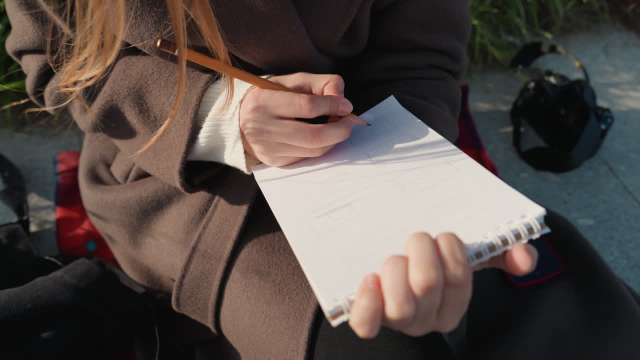 Giovane donna bianca che scrive su un taccuino all'aperto, primo piano delle mani con la penna, cappotto di lana e borsa accanto al casco, luce solare soffusa e tonalità autunnali, stato d'animo riflessivo mentre abbozza appunti per studio e poesia.