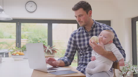 Working-father-using-laptop-at-home-whilst-feeding-baby-son-sitting-on-his-knee--from-bottle---shot-in-slow-motion