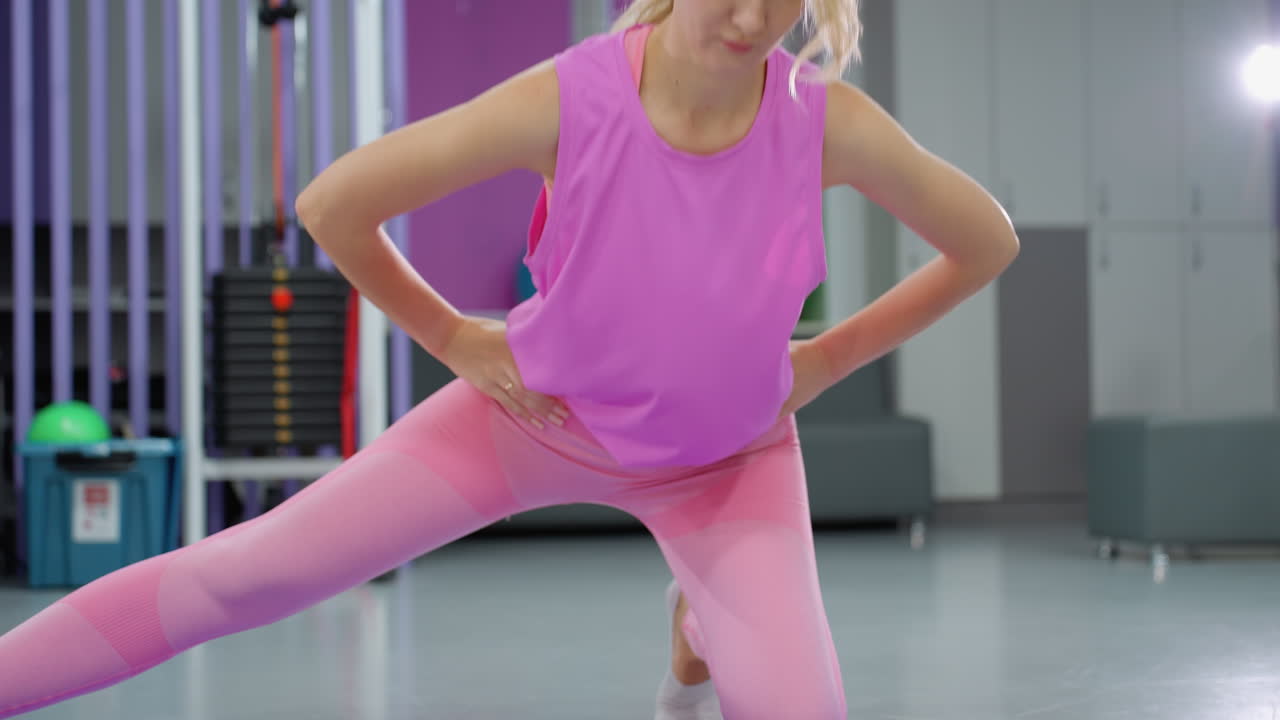 mujer con traje de entrenamiento rosa realizando estiramiento de piernas con las manos en la cintura, equilibrio mientras baja y regresa al gimnasio, el fondo incluye equipos de levantamiento de pesas, luz brillante y sofá de gimnasio