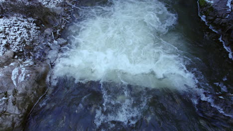 Primer-Plano-Aéreo-Cruzando-A-Lo-Largo-Del-Río-De-Montaña-Con-Una-Pequeña-Caída-De-Agua-Durante-El-Invierno-Con-Algo-De-Hielo-Y-Nieve,-Agua-Turbulenta,-Costa-Rocosa