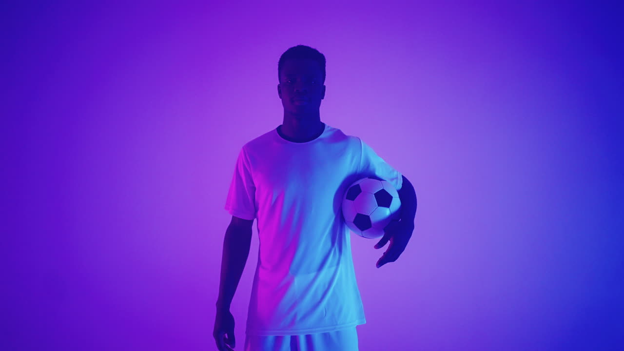 Ritratto al rallentatore di un giocatore di football americano professionista di colore in uniforme bianca, che guarda in camera sotto la luce neon blu-rossa dello studio. Un giocatore di football americano brutale, sicuro di sé e vittorioso