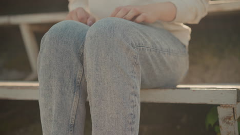 close-up of lady in jeans sitting on rustic stadium seating, hand resting on her leg while adjusting her position, warm sunlight highlights the worn texture of bench and denim