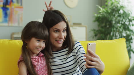 Primer-Plano-De-La-Madre-Y-La-Hija-Sonrientes-Tomando-Selfie-Divertido-En-El-Teléfono-Inteligente-En-El-Sofá-Amarillo