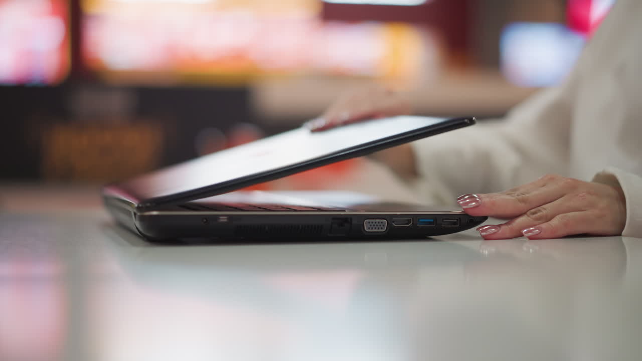 primer plano de manos elegantes con uñas pintadas que alcanzan una computadora portátil cerrada la abre en un entorno interior moderno, el fondo presenta una iluminación suave y borrosa con una sutil luz roja sonora