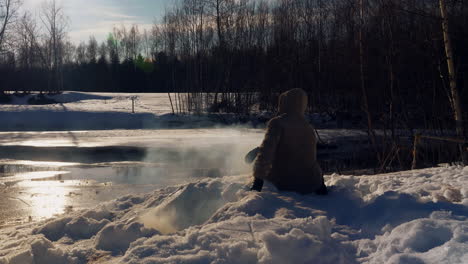 Arctic-Viking-settler-sitting-by-the-camp-fire,-icy-winter-lake-and-snowy-woodland-wilderness