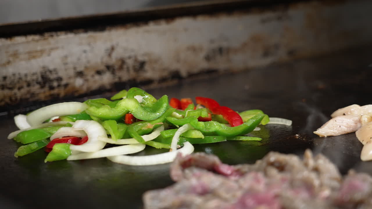 mano con guante añade rebanadas pimientos rojos verdes y cebollas a la parrilla plana caliente humeante en la cocina de un restaurante comercial como carne de pollo fajitas chisporrotean en primer plano, cámara lenta deslizador de primer plano 4k