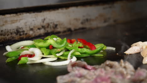 gloved hand adds sliced red green bell peppers and onions to steaming hot flat top grill in commercial restaurant kitchen as beef chicken fajitas sizzle in foreground, slow motion close up slider 4k