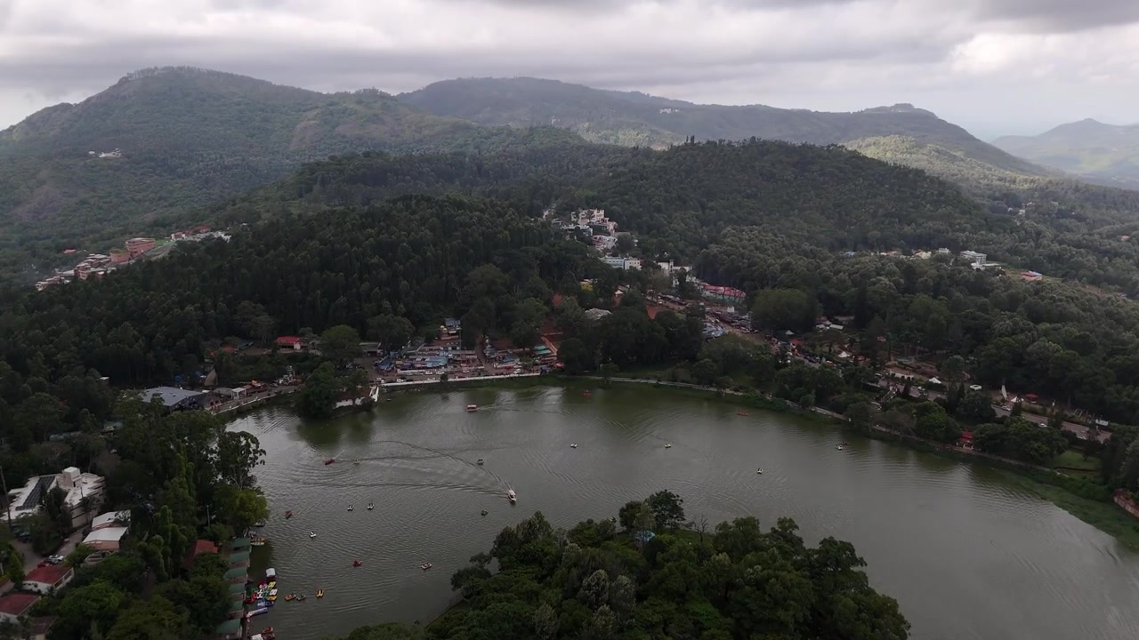 Yercaud Lake with its garden with a fountain and seating areas lines the water's edge. In the background, the town of Yercaud sprawls across rolling hills dotted with residential buildings