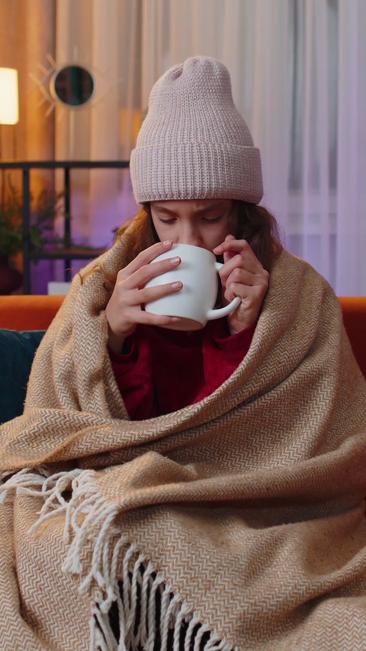 Ragazza adolescente malata, con un cappello e avvolta in un plaid, seduta sul divano, trema per il freddo bevendo cacao caldo.