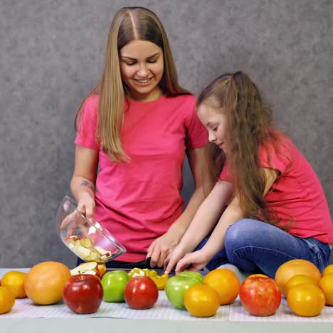 mother with daughter with fruit in the kitchen. young woman and a little child in pink t-shirts prepare fruit salad at home. healthy food.