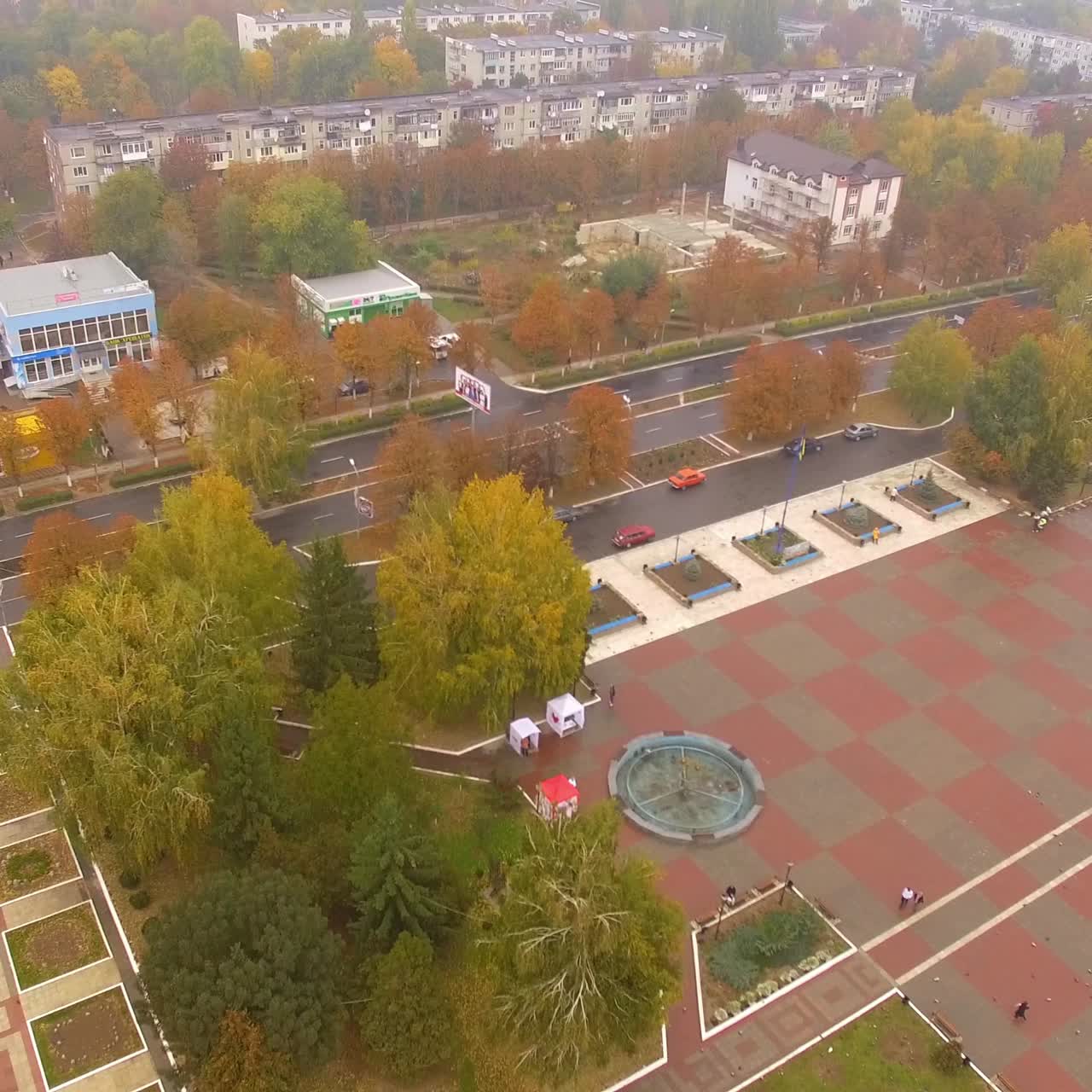 City square in downtown. Trees, fir-trees and flowerbeds around the area. Drone footage going up gradually over the cityscape