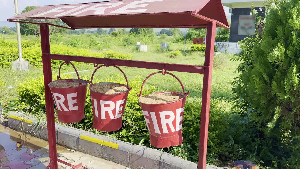 Three red fire buckets filled with sand hang from a metal stand in an outdoor area. A simple, traditional fire safety setup used in public places and industrial areas