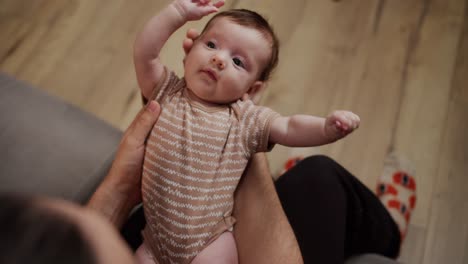 Close-up-from-above-a-happy-father-holds-his-little-baby-daughter-in-a-brown-overalls-in-his-arms-in-a-modern-apartment-at-home