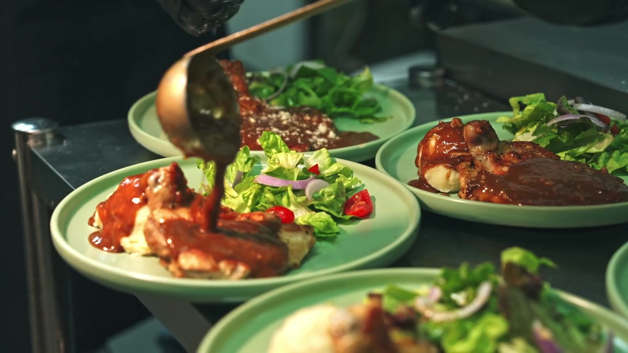 Medium tracking shot showing a kitchen line where a chef ladles hot sauce and garnishes grated cheese over multiple round plates of grilled chicken chops.