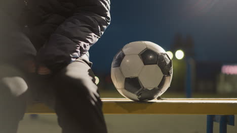 close up of someone seated on a bench with a worn soccer ball beside them, illuminated by the dim night lights in the background, the ball slightly moves with the wind