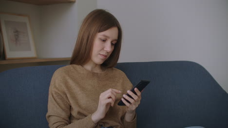 Mujer-Joven-Sentada-En-El-Escritorio-De-La-Oficina-En-Casa-Distraída-Del-Trabajo-Sosteniendo-Un-Teléfono-Inteligente-Usando-Un-Sitio-Web-De-Redes-Sociales-Charlando-Con-Un-Amigo-De-Forma-Remota-Usando-Un-Dispositivo-Moderno-Y-Una-Conexión-A-Internet