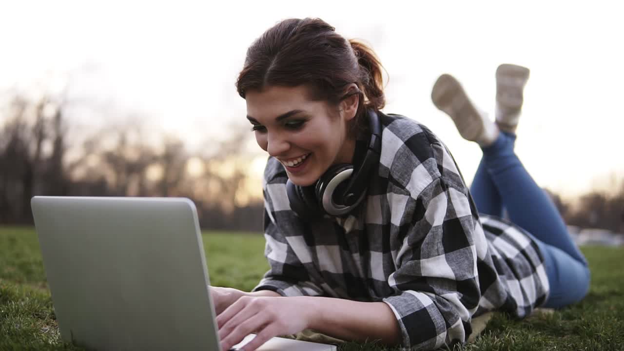 hermosa chica yace en la hierba en el parque. utiliza una computadora portátil, charlando. auriculares en el cuello. ella tiene un buen tiempo en la comunicación. sonrisas, feliz. día brillante