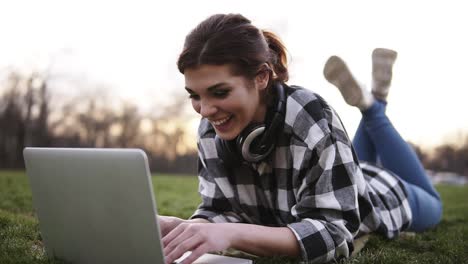 Schönes-Mädchen-Liegt-Auf-Dem-Gras-Im-Park.-Benutzt-Einen-Laptop-Und-Chattet.-Kopfhörer-Am-Hals.-Sie-Hat-Spaß-An-Der-Kommunikation.-Lächelt,-Glücklich.-Heller-Tag