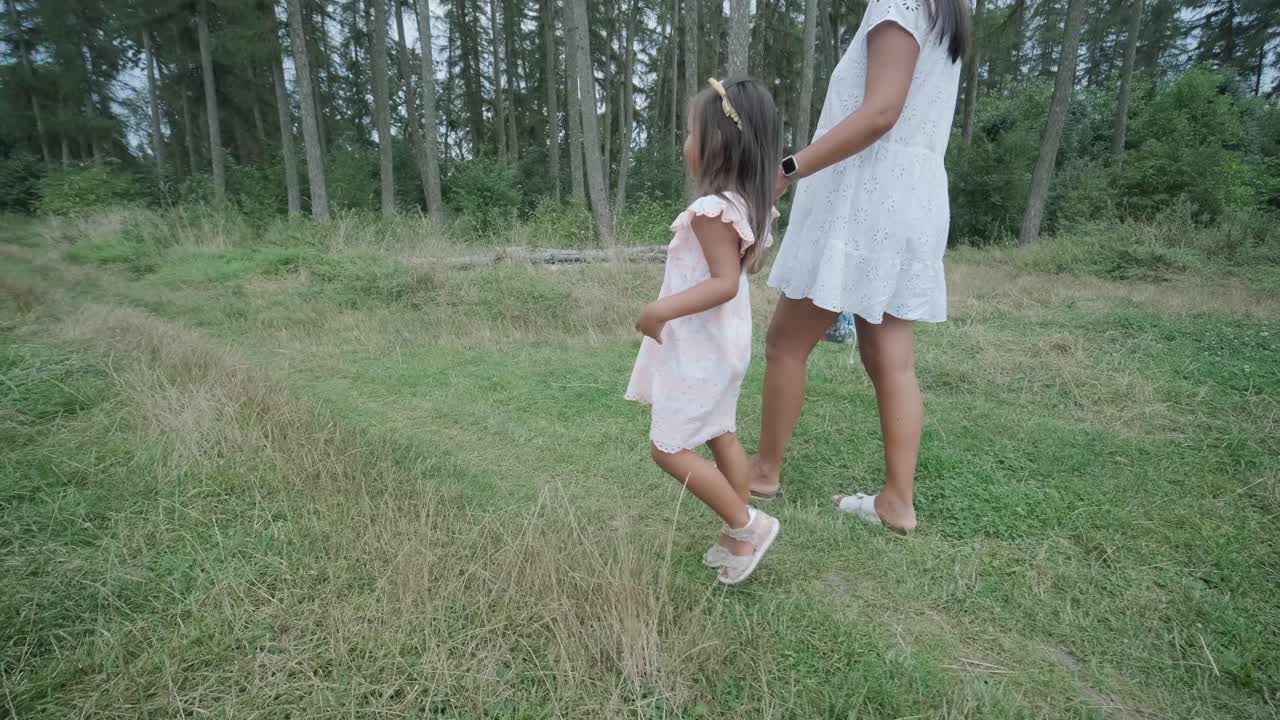 Una donna incinta e la sua giovane figlia, entrambe vestite di bianco, camminano mano nella mano attraverso un parco erboso. La scena cattura un momento sereno di legame familiare nella natura, circondate da alberi