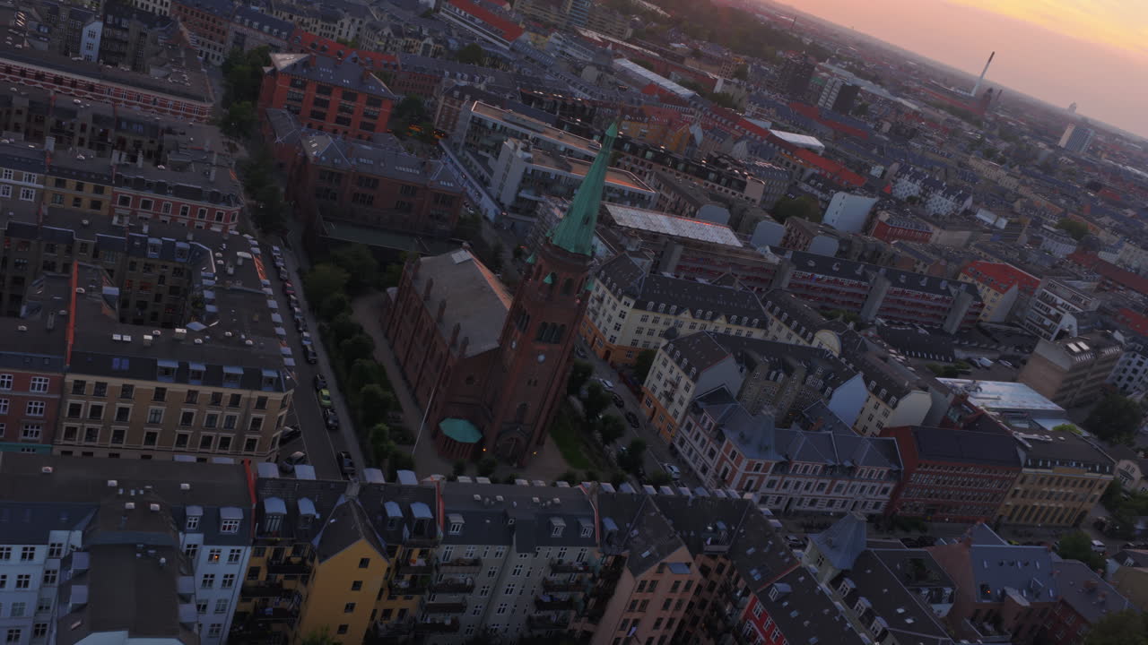 Aerial drone view of warm evening light over the rooftops of Vesterbro and Frederiksberg, with industrial chimneys and towers on the horizon in Copenhagen, Denmark