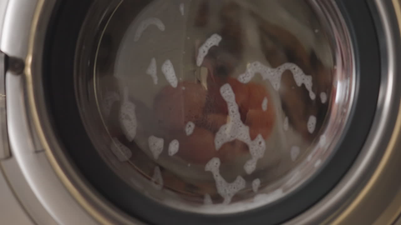 Close up on stainless washer number five showing clothes swirling behind glass door, soap suds and water bubbles circling during wash cycle inside bright laundromat, clean outcome, smooth drum motion