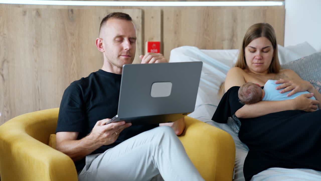 Husband finishes work at laptop and closes it. Man turns to his wife holding their newborn.