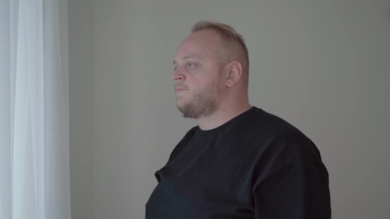 Middle aged man in a dark sweatshirt gazes out sheer curtains, quietly reflecting on a difficult life decision at home.