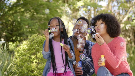 happy african american parents with daughter blowing bubbles in garden at home, slow motion