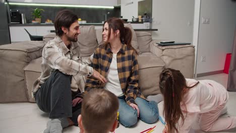 un hombre moreno con paja en una camiseta beige se comunica con su esposa en una camisa a cuadros sentado en el suelo mientras sus hijos están haciendo creatividad y dibujando en hojas de papel sentados en el suelo y divirtiéndose en un nuevo apartamento moderno