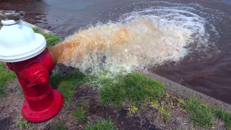 a low angle shot of a red and white fire hydrant spraying rusty water onto the street on a sunny day