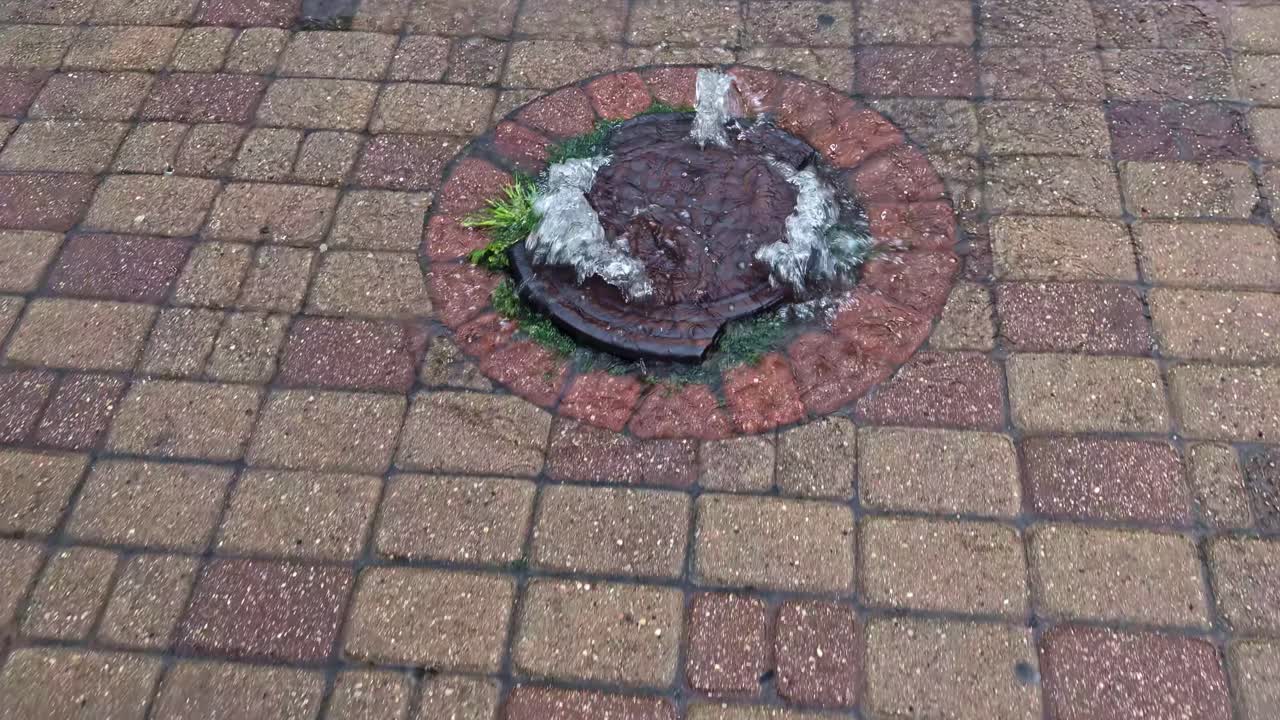 Water overflows from a clogged street manhole cover, bubbling up on a brick sidewalk on a rainy day as woman's foot steps on it