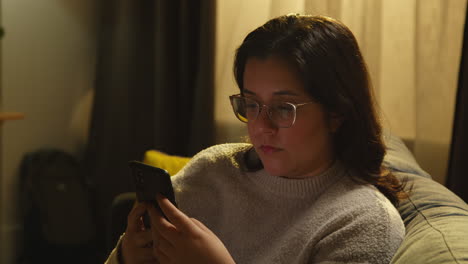 Young Woman At Home Sitting On Sofa With Mobile Phone Scrolling Through Internet Or Social Media In The Evening