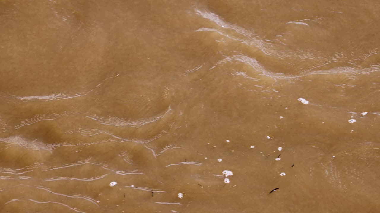 Drone captures muddy river water currents and foam patterns in daylight, steady overhead perspective