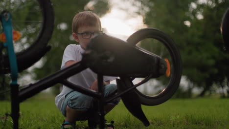 a young boy is sitting on the grass, intently rotating the pedals of his bicycle, causing the tire to spin, he appears focused, as his younger brother stands nearby, watching him