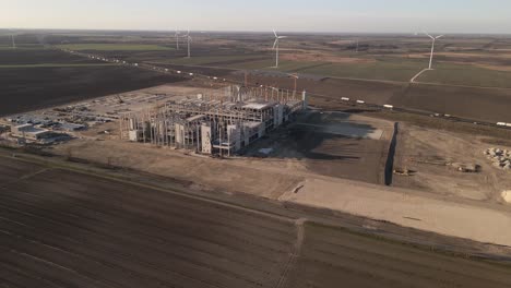an aerial view captures an industrial construction site in a rural area, featuring cranes, partially built structures, and construction equipment. next to a busy highway with moving trucks and cars.