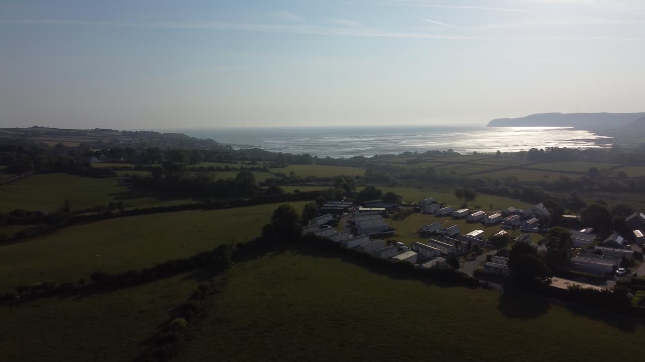 Aerial view Welsh caravan park at sunrise overlooking shimmering coastal bay and rolling countryside hills, panning left