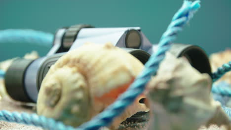 close-up product video of blue and black binoculars with sea shells and blue rope, surrounded by beach sand, on a turntable display