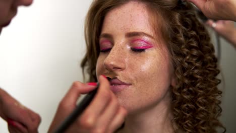 Unrecognizable-makeup-artist-applies-toner-on-the-face-of-a-beautiful-model-with-freckles,-red-headed.-Hairdresser-with-a-curling-iron-making-hair-style-in-tandem.-Close-up.-Slow-motion