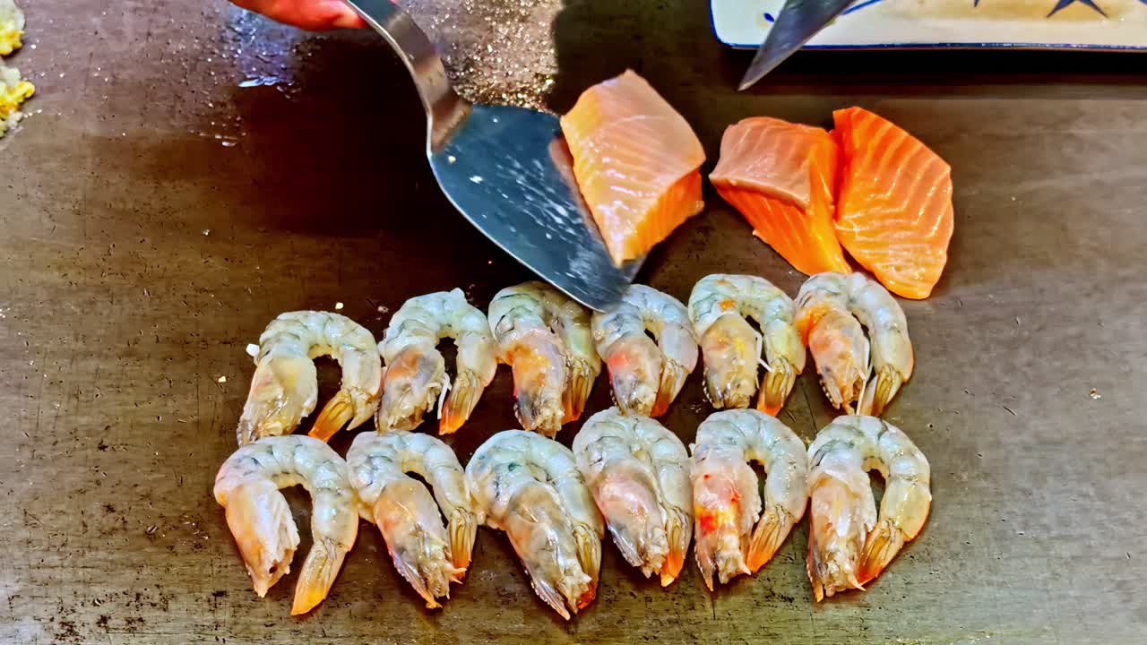 Chef arranging salmon and shrimp fillets side by side on flat top grill in restaurant kitchen, cooking heat