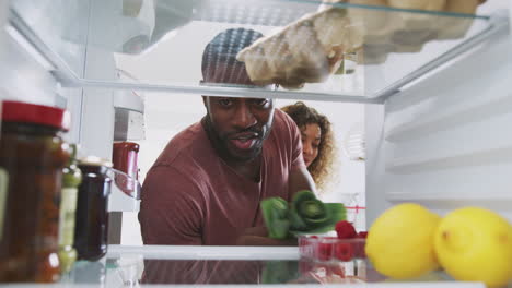 view looking out from inside of refrigerator as couple open door and unpacks shopping bag of food