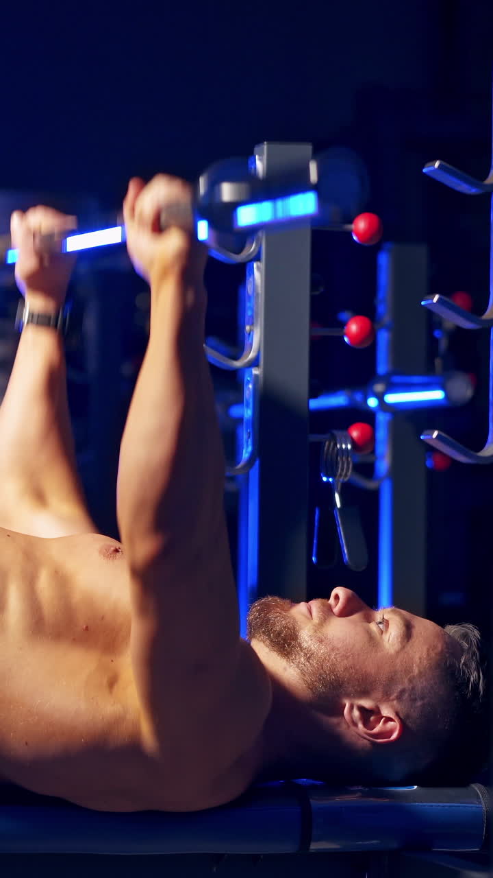 Shirtless young bodybuilder doing bench press for chest. Strong abs. Blue light filter. Video from the side. Vertical video