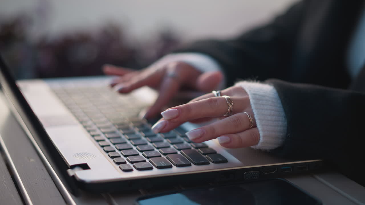 Close-up of hands with elegant manicured nails typing on laptop keyboard outdoors, wearing rings and a cozy sweater sleeve, with blur flower nearby, reflection visible on phone placed on table