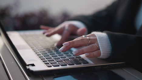 close-up of hands with elegant manicured nails typing on laptop keyboard outdoors, wearing rings and a cozy sweater sleeve, with blur flower nearby, reflection visible on phone placed on table