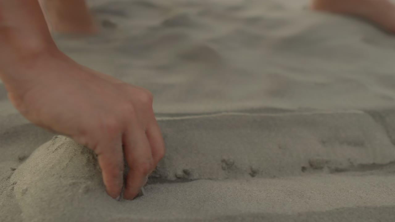The camera focuses closely on a woman's hands as she delicately picks up a seashell from the sandy beach capturing a moment of serene beachcombing
