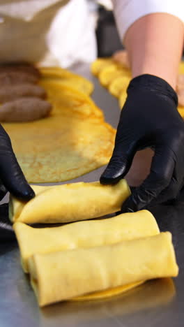 the cook in black gloves rolling meat into yellow crepes. process of making stuffed pancakes at modern restaurant/food factory. vertical video